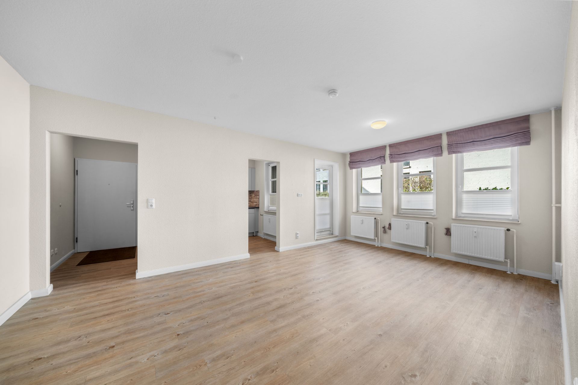Helles Wohnzimmer mit Holzboden, drei Fenstern und Zugang zur Küche.