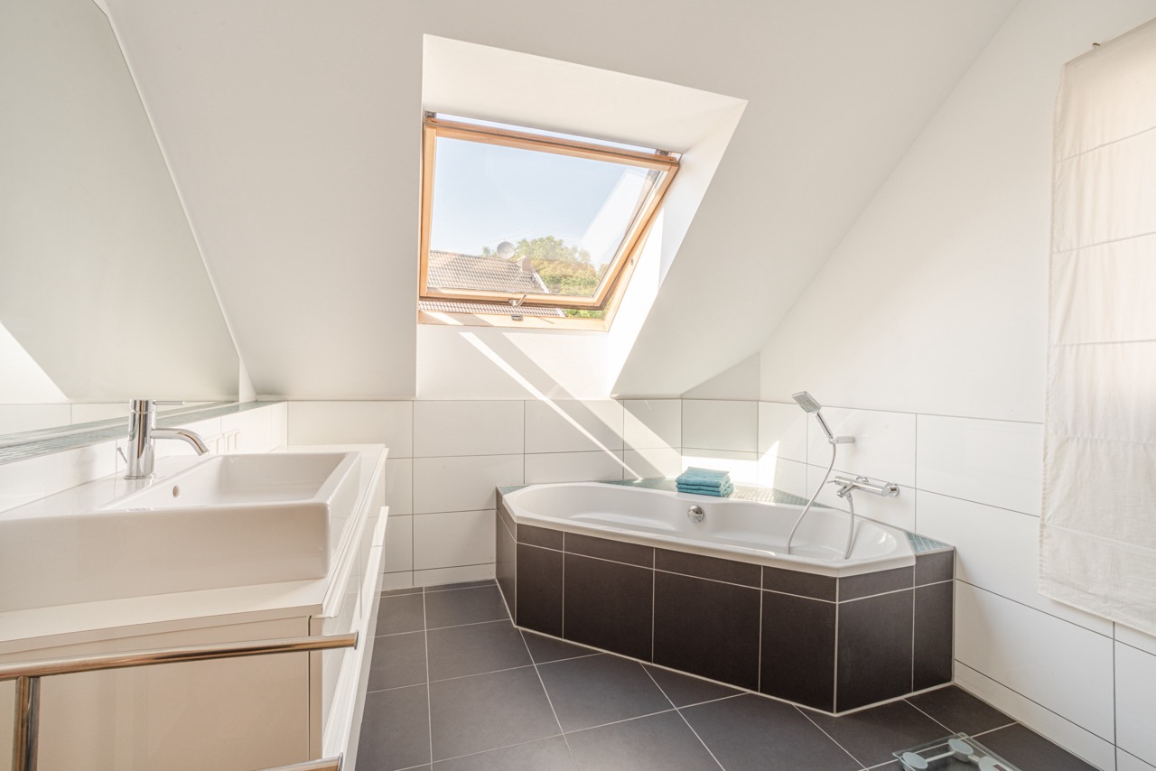 Helles Bad mit Eckbadewanne, Fenster im Dach, Waschbecken und weißen Fliesen.