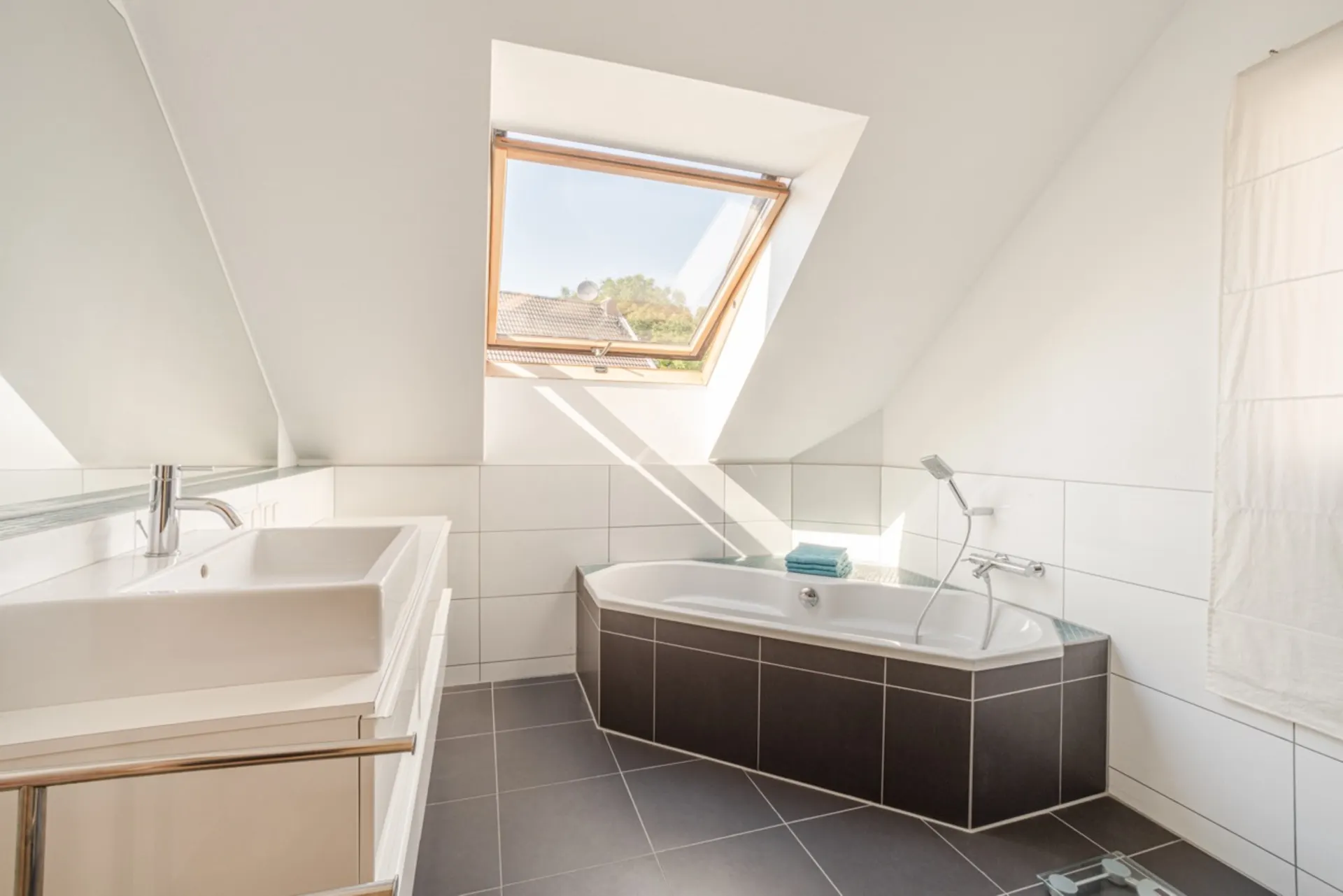 Helles Bad mit Eckbadewanne, Fenster im Dach, Waschbecken und weißen Fliesen.