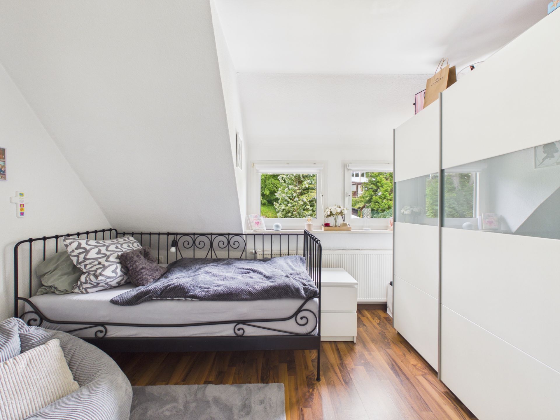 Helles Schlafzimmer mit einem Einzelbett, weißem Schrank, zwei Fenstern und Holzboden.