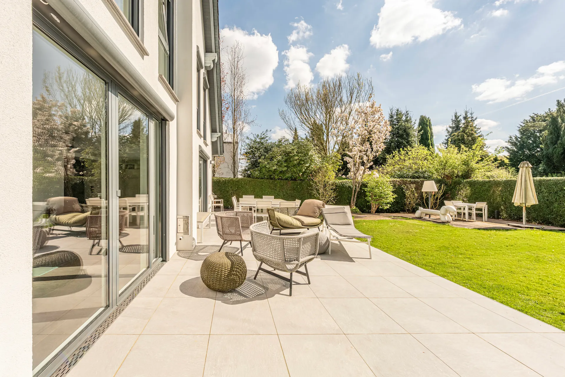 Moderne Terrasse mit Sitzmöbeln am Haus, daneben grüner Garten mit Bäumen und Gartenmöbeln.