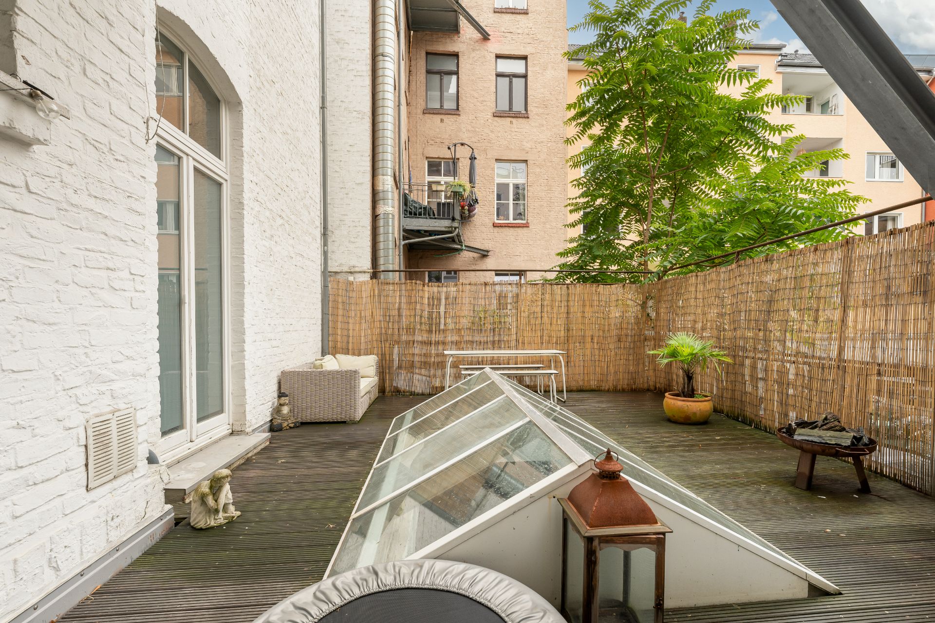 Große Holzterrasse mit Pflanzkübel, Sitzbank, Sofa und Glasfenster, umgeben von einem Sichtschutzzaun.