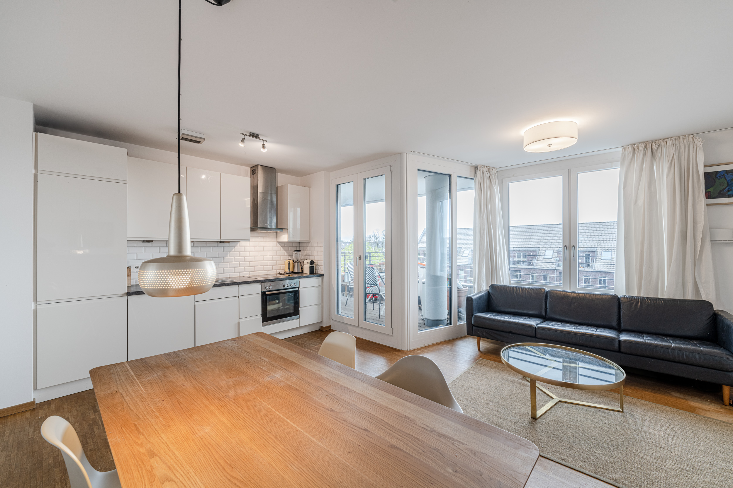 Helles Wohnzimmer mit offener Küche, Esstisch, schwarzem Sofa und Zugang zu einem Balkon.