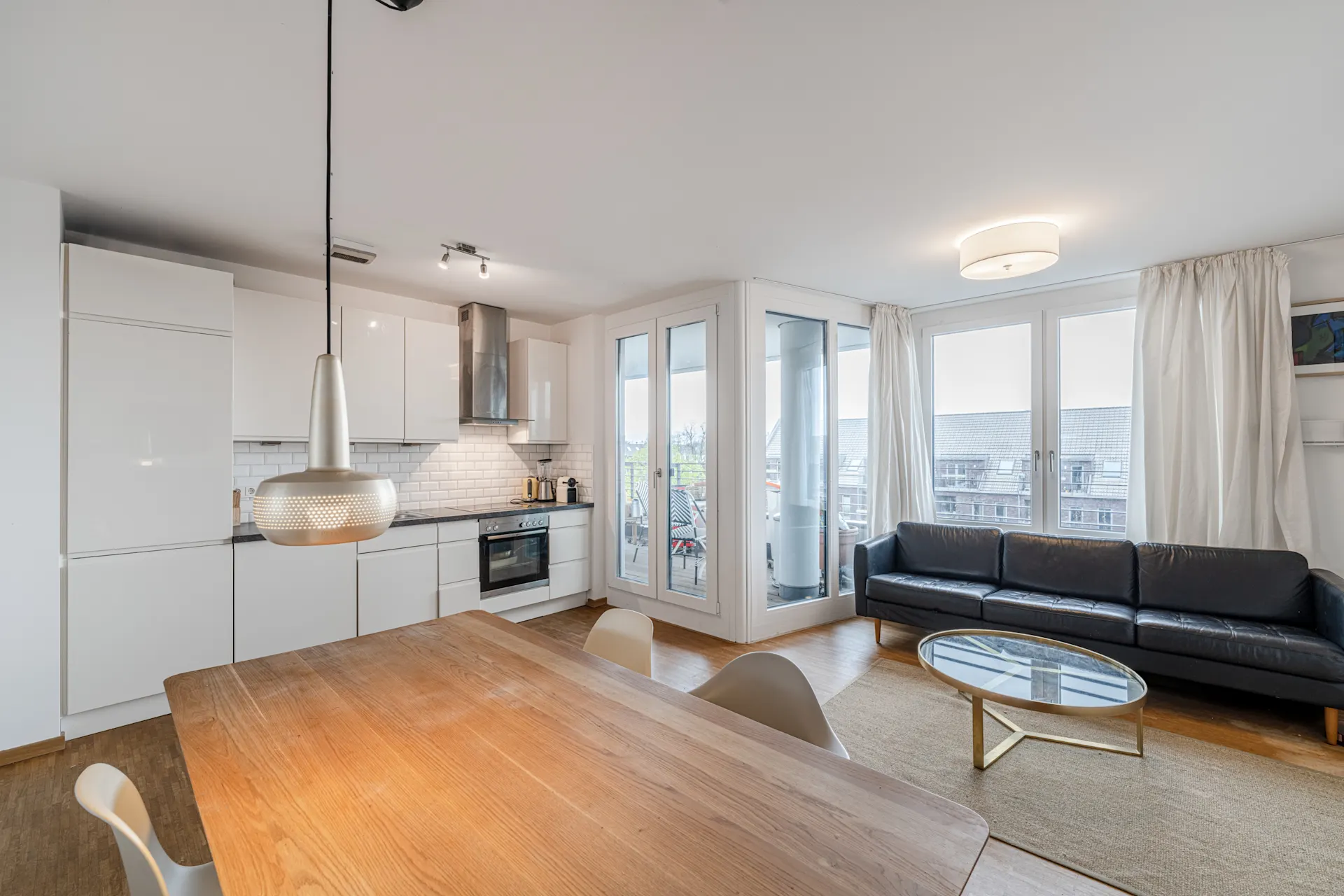 Helles Wohnzimmer mit offener Küche, Esstisch, schwarzem Sofa und Zugang zu einem Balkon.