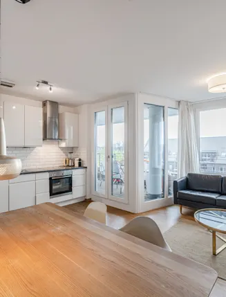 Helles Wohnzimmer mit offener Küche, Esstisch, schwarzem Sofa und Zugang zu einem Balkon.