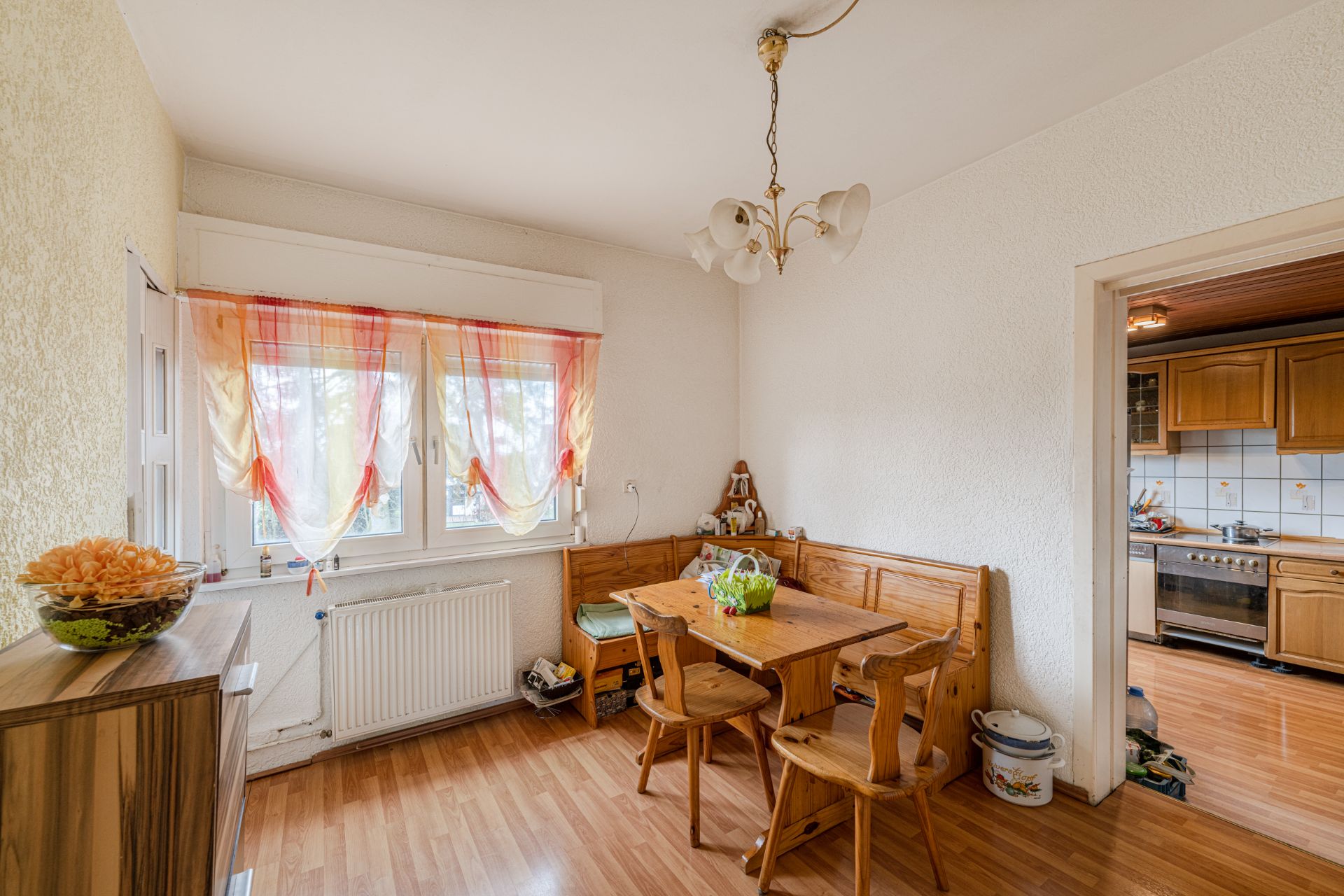 Essbereich mit Holztisch, Stühlen und Fenster mit bunten Vorhängen, daneben eine offene Küche.
