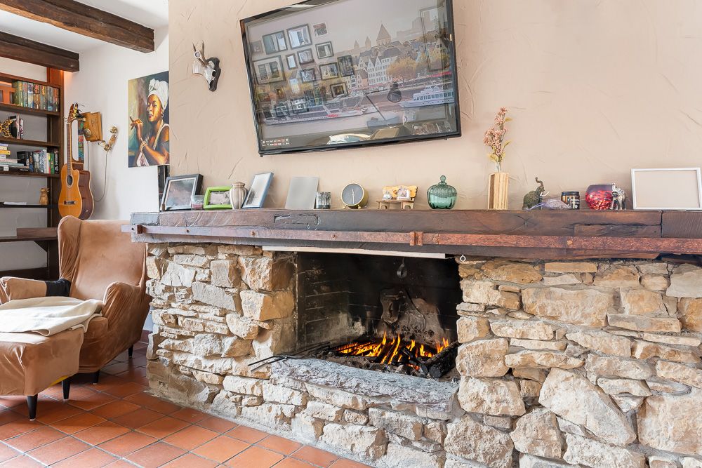 Wohnzimmer mit offenem Steinkamin, brennendem Feuer, Sessel und Regal mit Büchern und Gitarre.