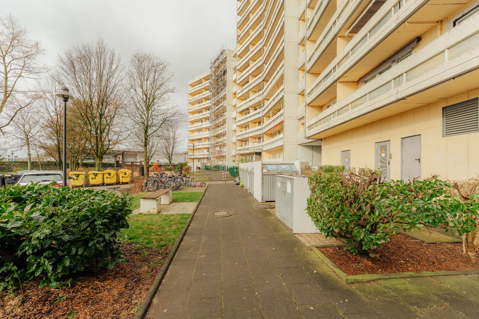 Gehweg mit Büschen und Bänken vor einem mehrstöckigen Wohnhaus.