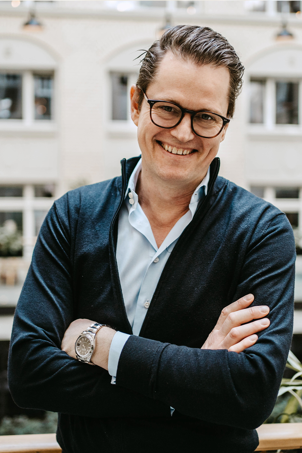 Ein Mann mit Brille und Uhr steht lächelnd mit verschränkten Armen vor Fenstern.