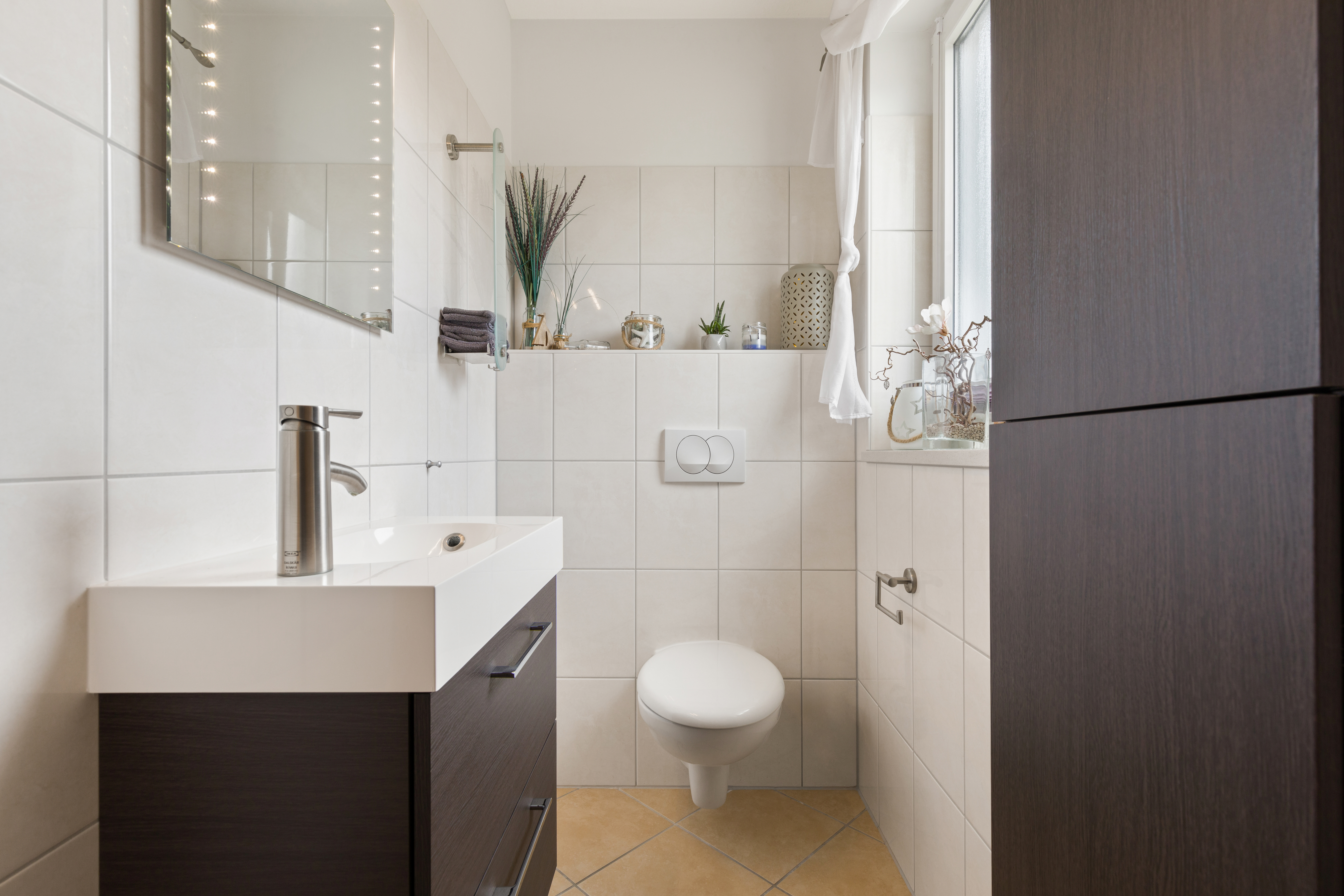 Modernes Badezimmer mit Wandtoilette, Waschbecken und Fenster, helle Fliesen und Dekorationen.