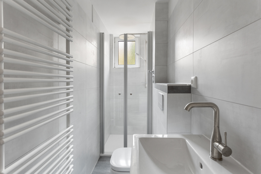 Modernes, schmales Badezimmer mit Dusche, Waschbecken, Toilette und Handtuchheizkörper.