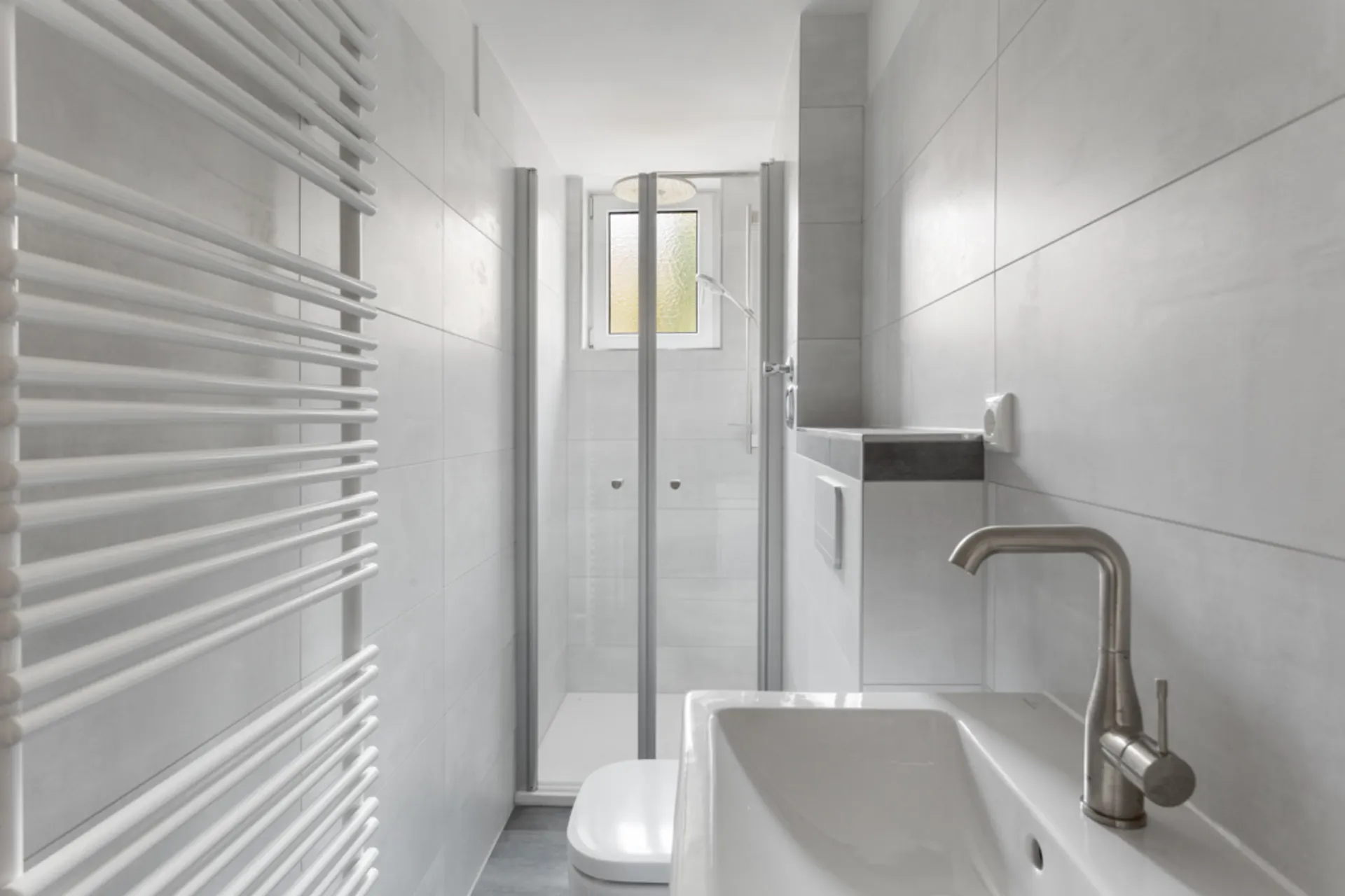 Modernes, schmales Badezimmer mit Dusche, Waschbecken, Toilette und Handtuchheizkörper.