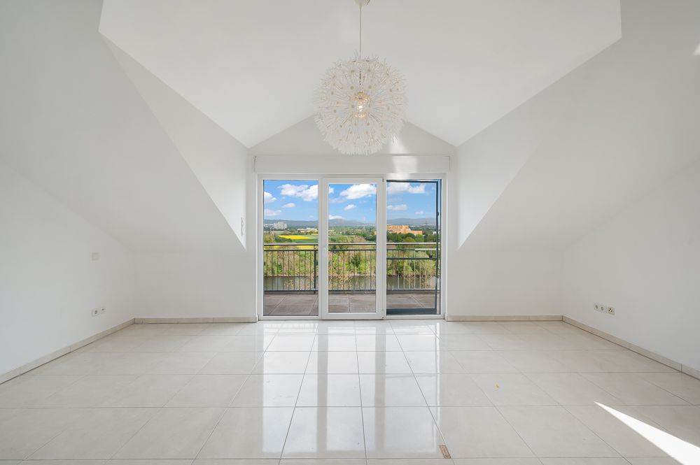 Heller, leerer Raum mit Fliesenboden, großer Fensterfront und Blick auf einen Balkon mit Landschaft dahinter.