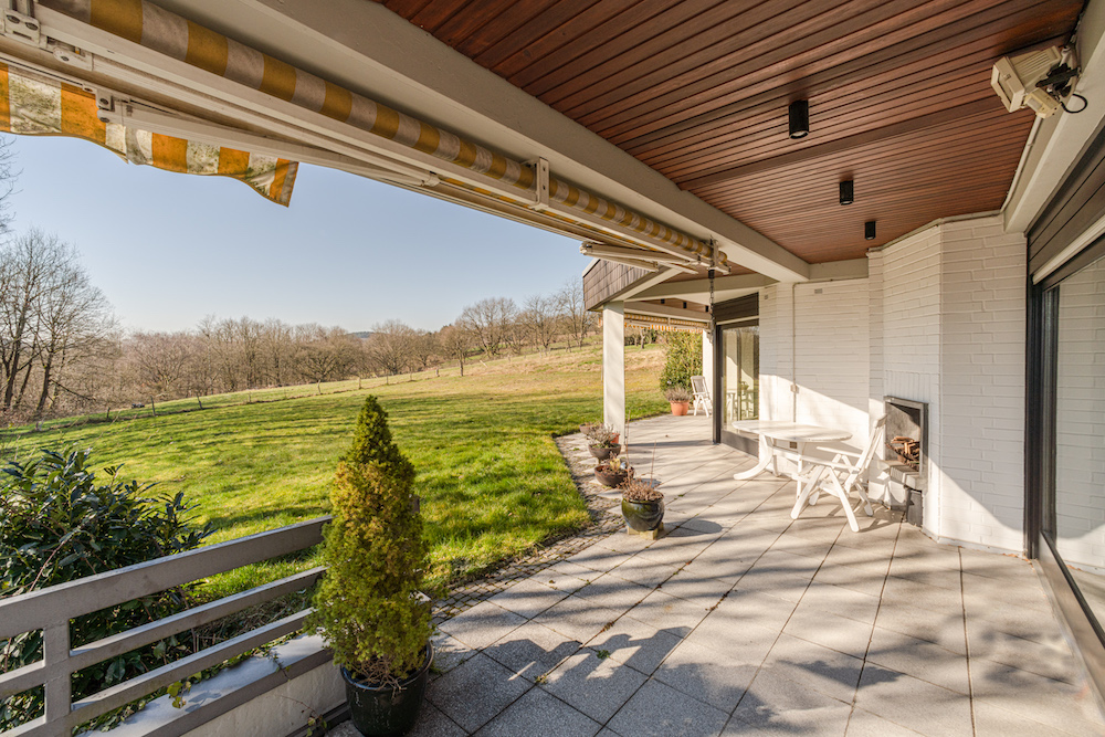 Überdachte Terrasse mit Gartentisch, Stühlen und Blick auf eine große Wiese und Bäume.
