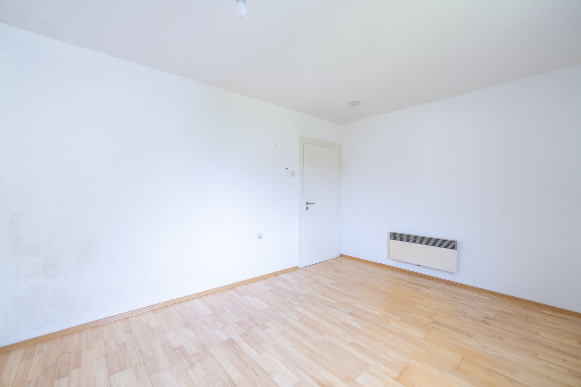Leerer, heller Raum mit Holzboden, weißen Wänden, Tür und Wandheizung.