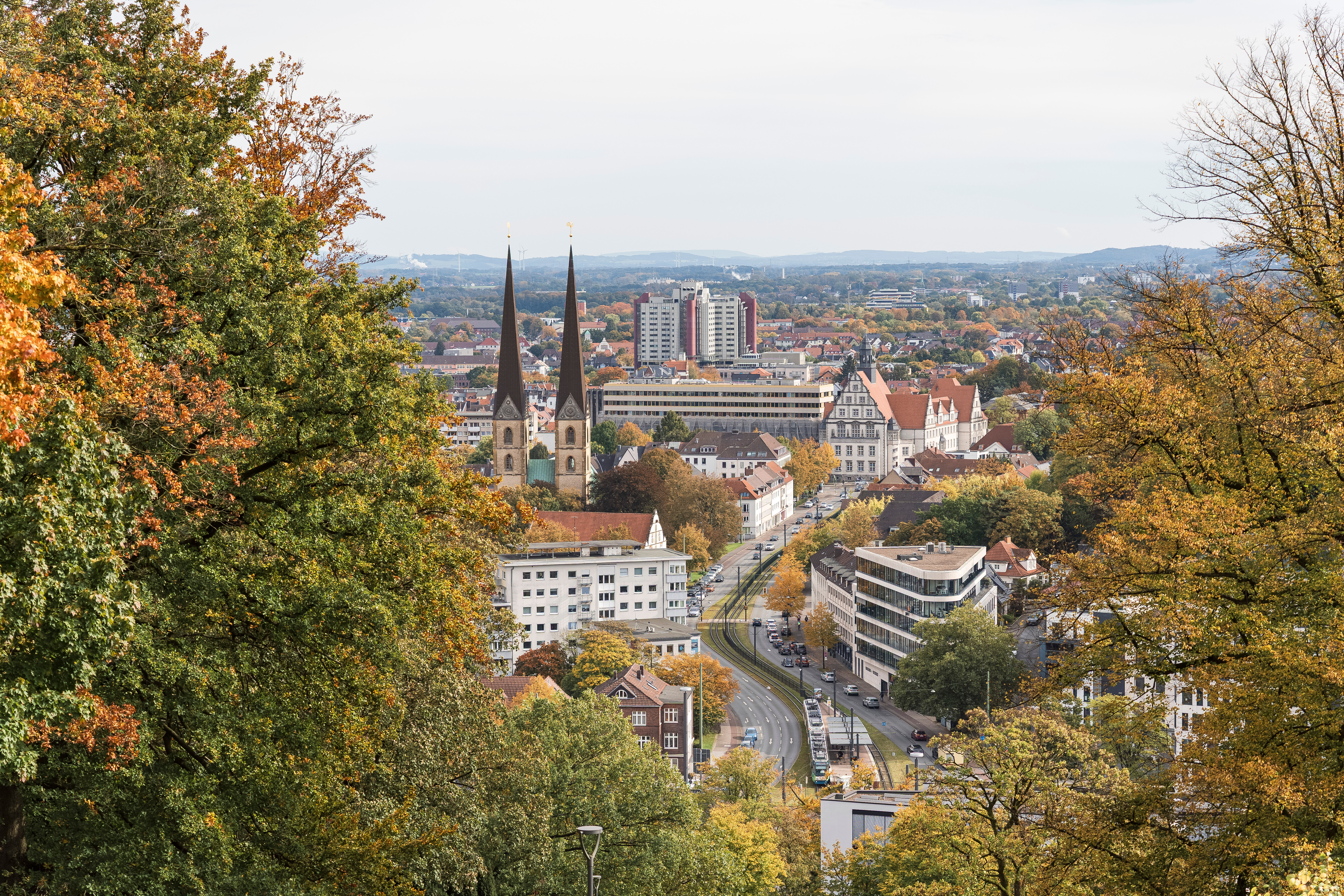 Stadtansicht mit zwei Kirchtürmen, mehreren Gebäuden und herbstlichen Bäumen im Vordergrund.