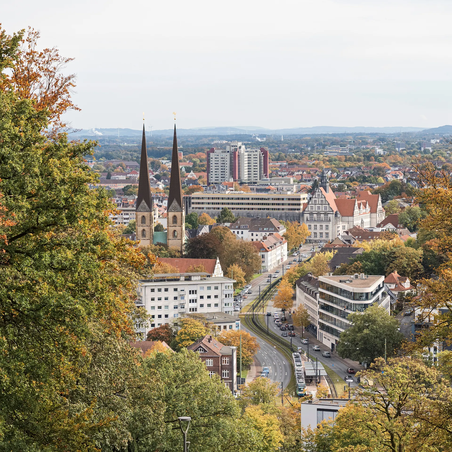 Stadtansicht mit zwei Kirchtürmen, mehreren Gebäuden und herbstlichen Bäumen im Vordergrund.