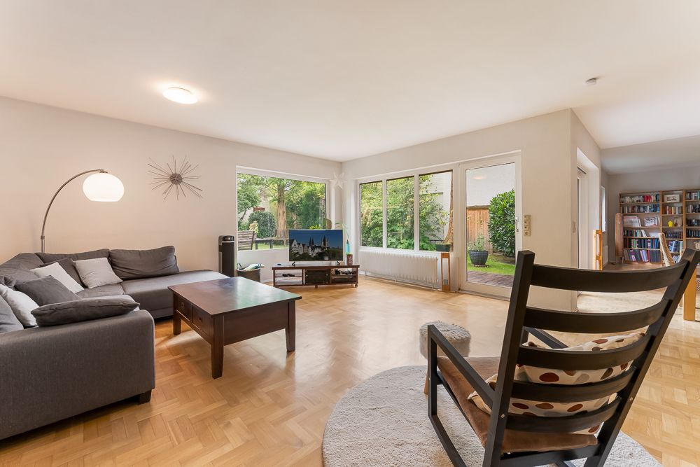 Helles Wohnzimmer mit Sofa, Sessel, Couchtisch, Fernseher und großen Fenstern zum Garten.