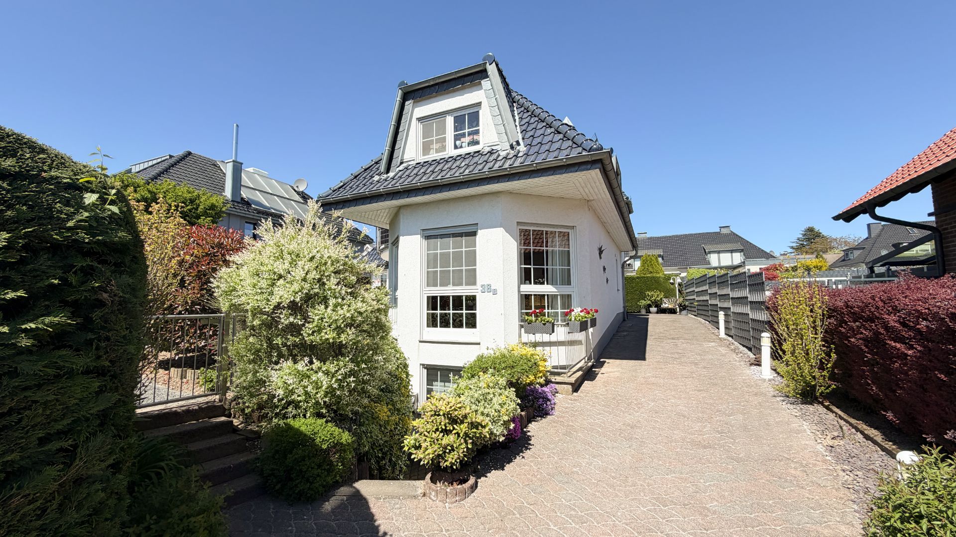 Weißes Wohnhaus mit dunkelgrauem Dach, Vorgarten und gepflasterter Einfahrt bei Sonnenschein.