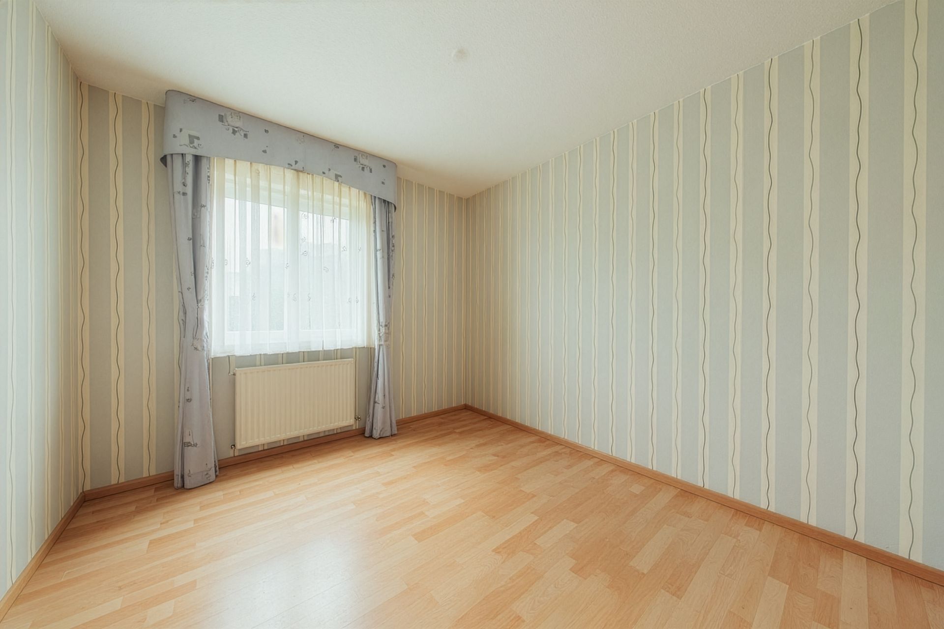 Leerer Raum mit Holzfußboden, gestreifter Tapete und Fenster mit Vorhang.