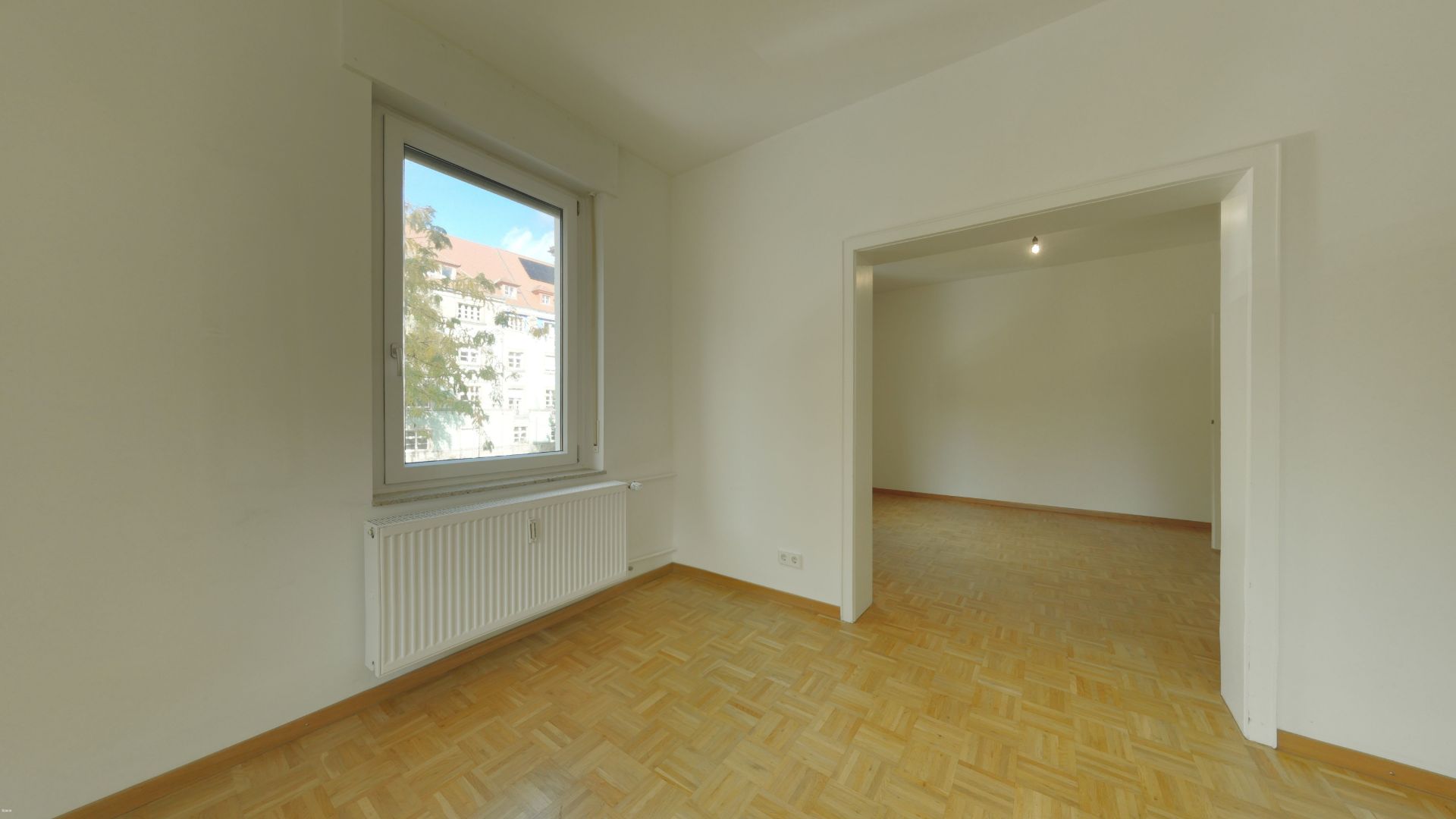 Helles Zimmer mit Parkettboden, Fenster und offenem Durchgang zu einem weiteren Raum.