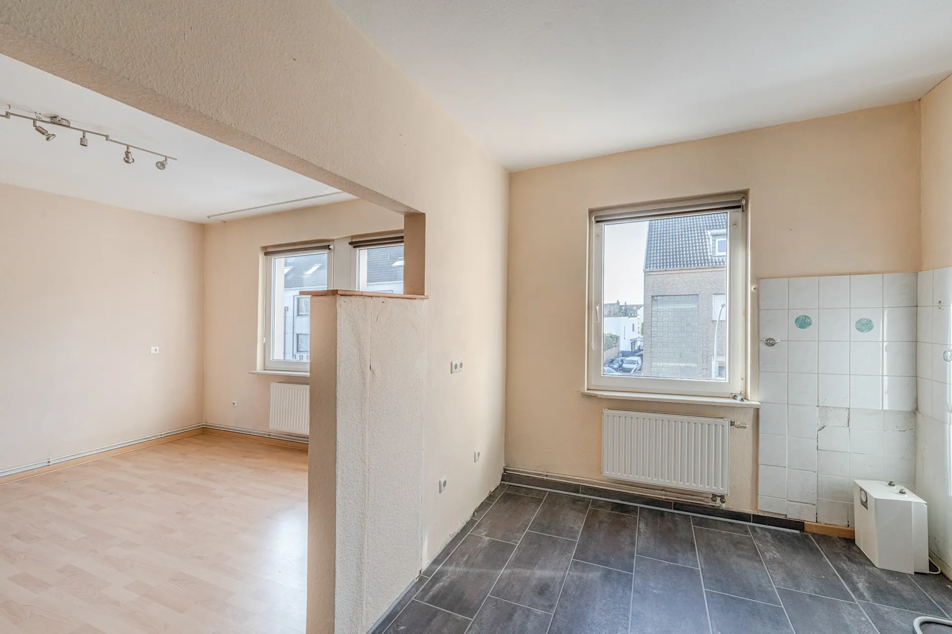 Leerer Wohnraum mit zwei großen Fenstern, hellem Boden und gefliester Ecke.
