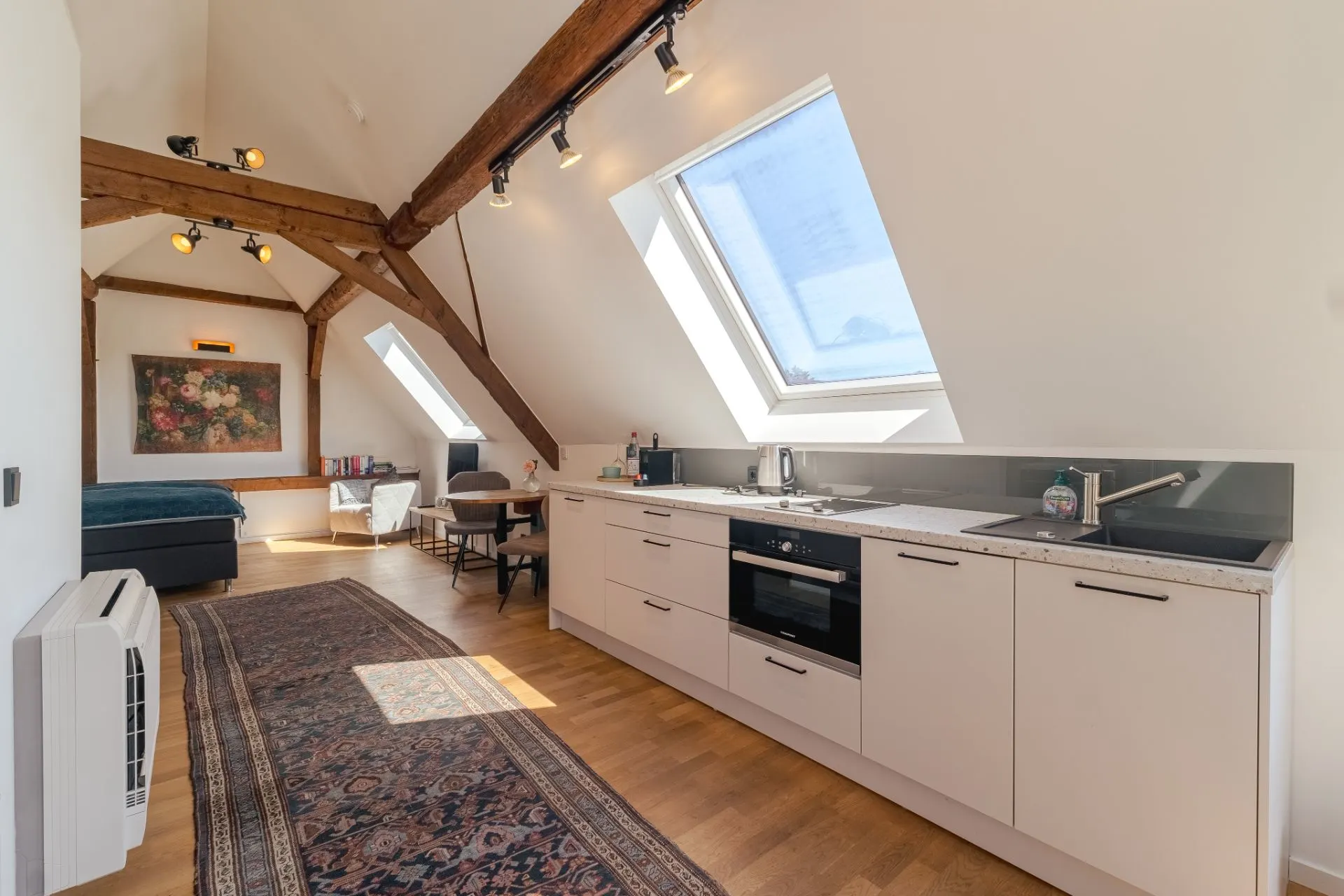 Helle Dachgeschosswohnung mit Holzbalken, Einbauküche, Esstisch und sichtbarem Bett.
