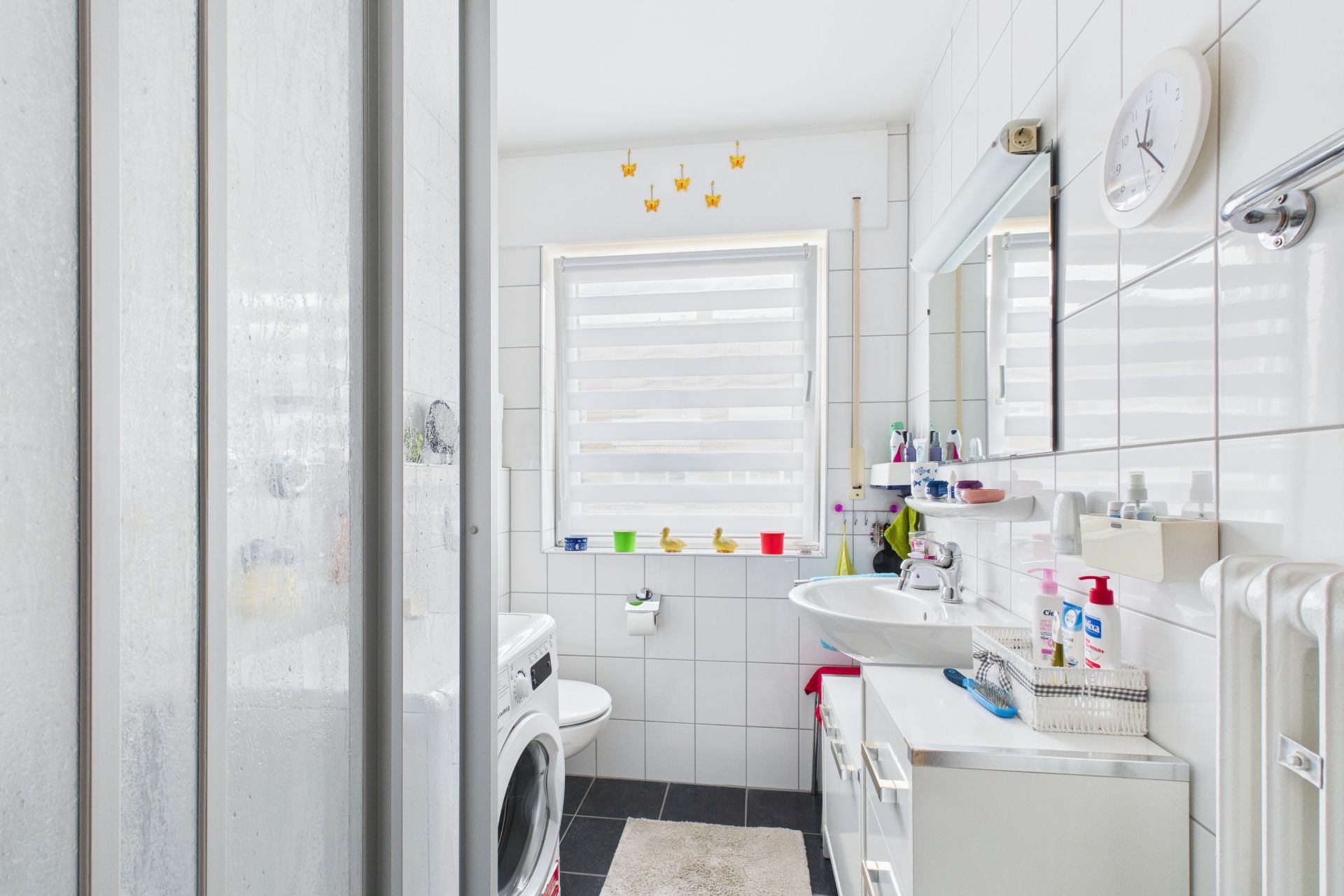 Modernes, helles Badezimmer mit Dusche, Waschmaschine, Waschbecken und Fenster mit Jalousie.