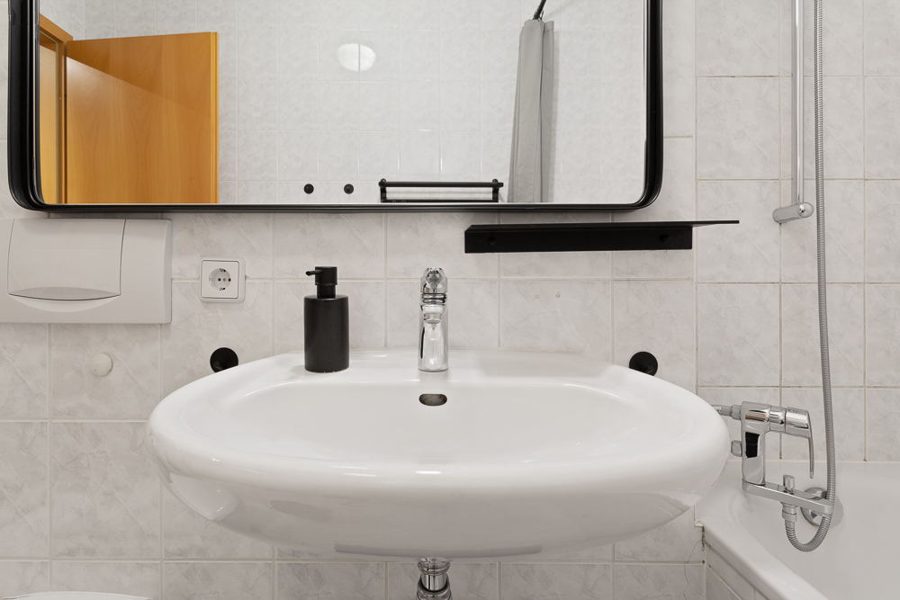 Waschbecken mit Spiegel, Seifenspender und Duscharmatur in einem weißen Badezimmer.