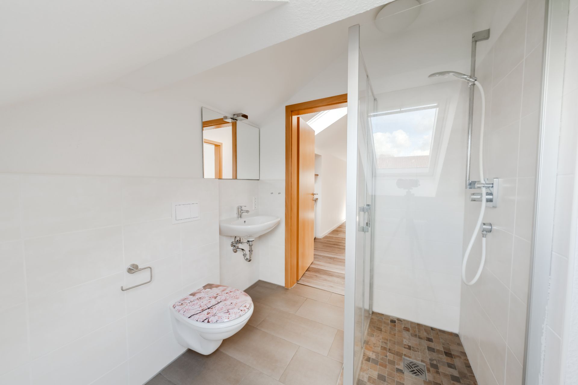 Modernes Badezimmer mit Dusche, Wandtoilette, Waschbecken und Spiegel unter einem Dachfenster.