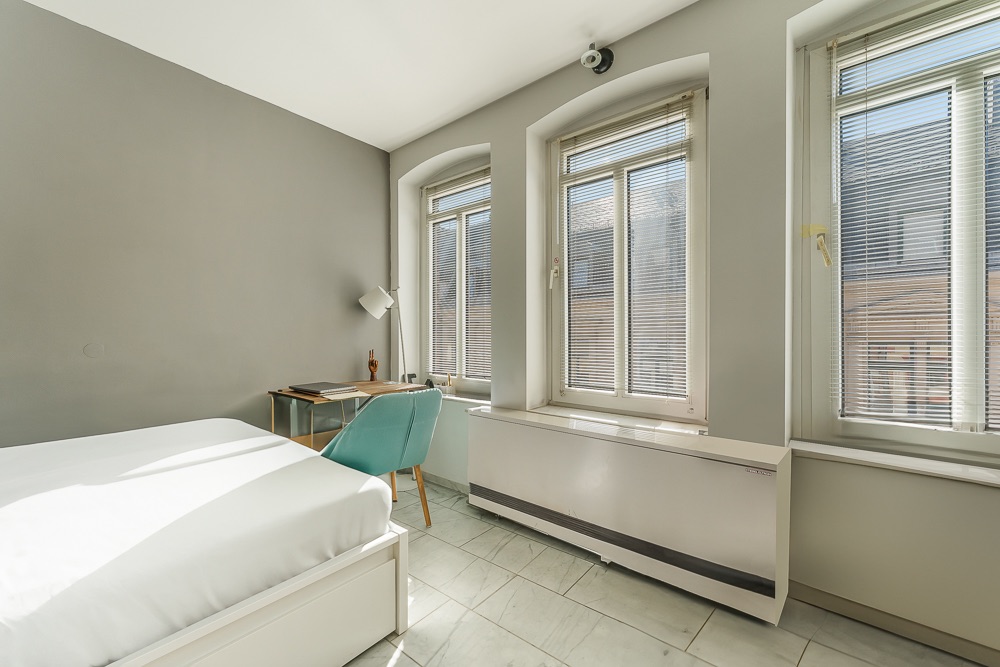 Helles Schlafzimmer mit Bett, Schreibtisch und drei großen Fenstern mit Jalousien.