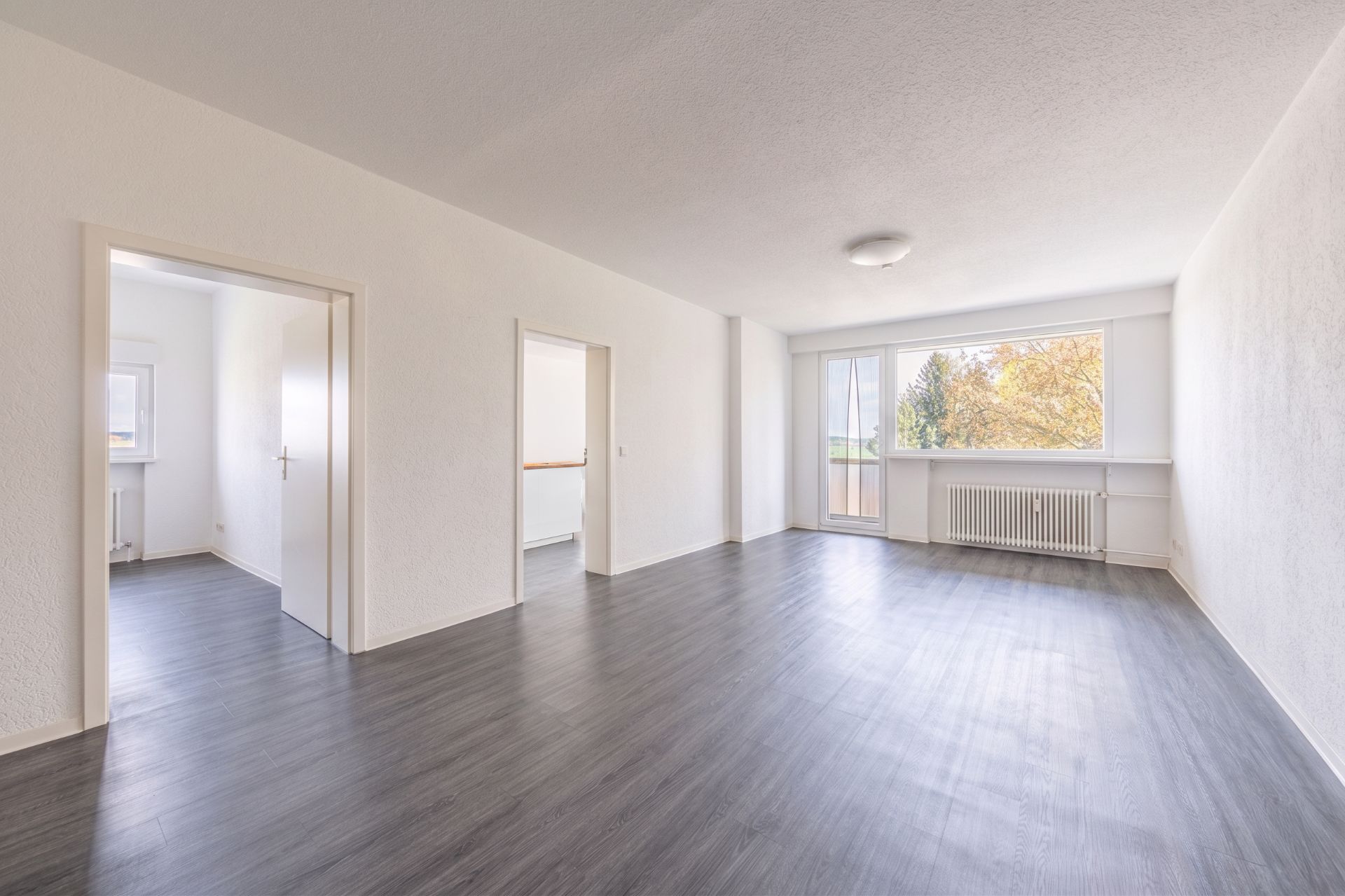 Helles, leeres Wohnzimmer mit großem Fenster, Tür zum Balkon und dunklem Boden.