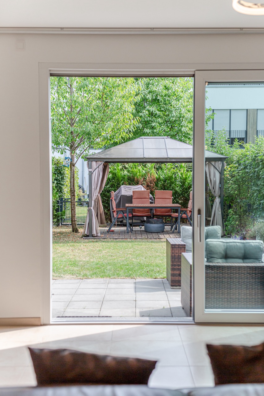 Blick durch eine Glastür auf eine Terrasse mit Sitzgruppe und Pavillon im Garten.