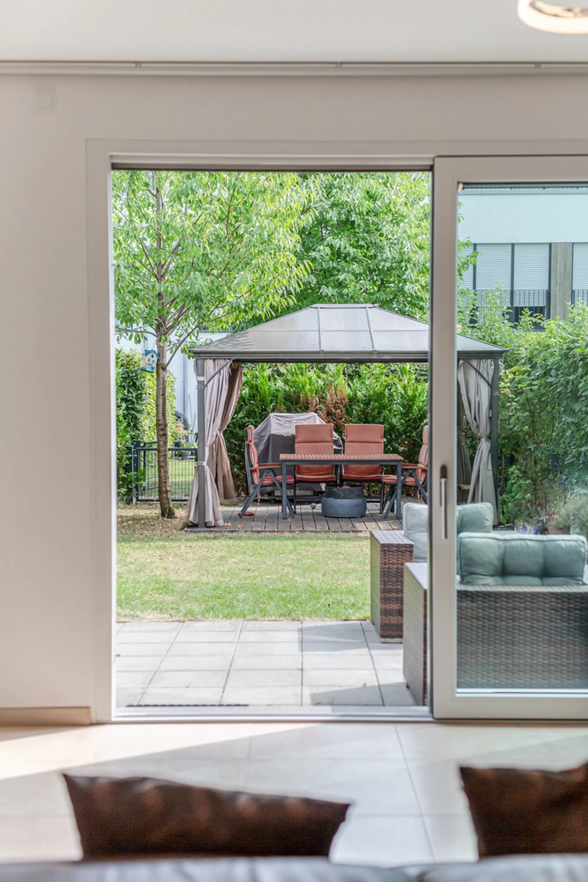 Blick durch eine Glastür auf eine Terrasse mit Sitzgruppe und Pavillon im Garten.