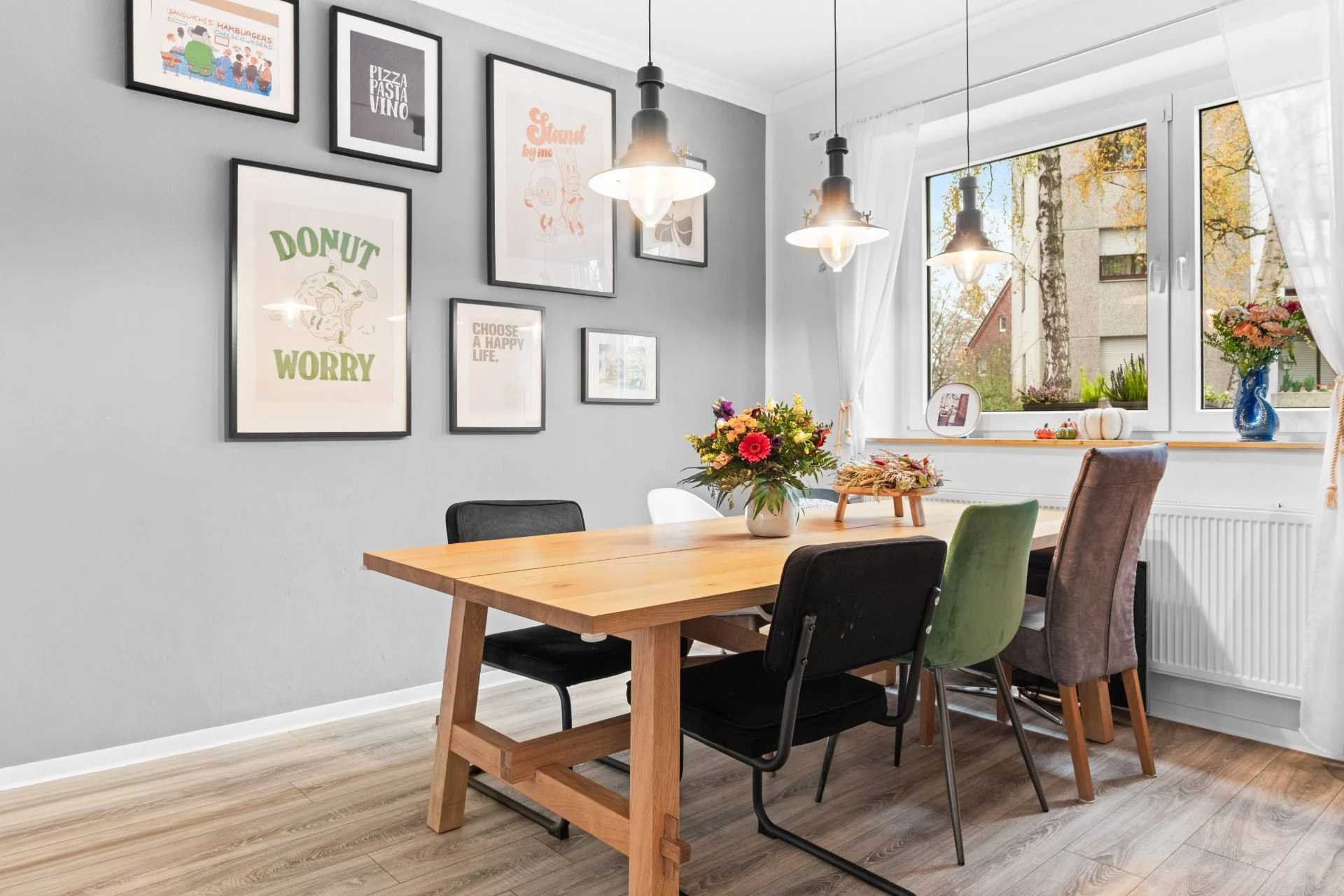 Essbereich mit Holztisch, verschiedenen Stühlen, Wandbildern und großem Fenster mit Blumenvasen.