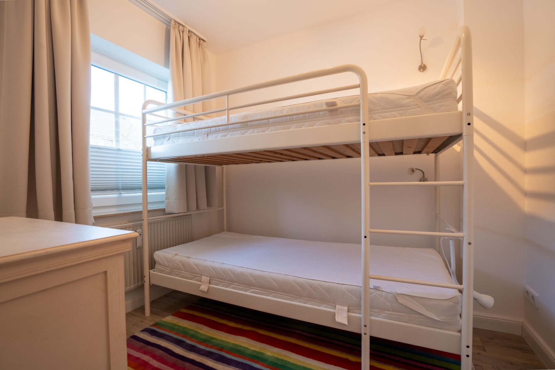 Zimmer mit Etagenbett, bunter Teppich, Fenster mit Vorhängen, weiße Wände und Kommode.