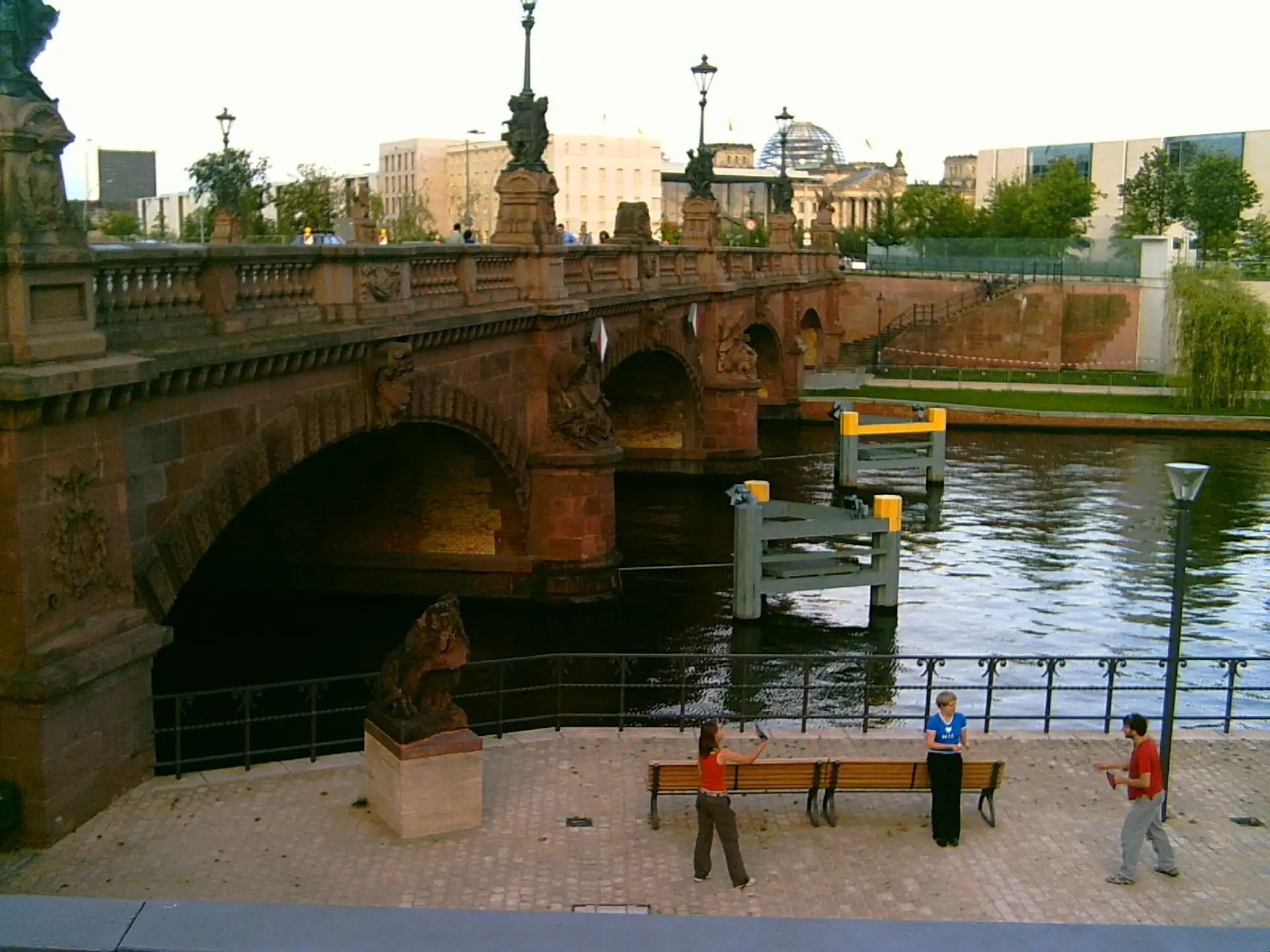 Drei Personen stehen auf einem gepflasterten Platz am Fluss unter einer steinernen Brücke.