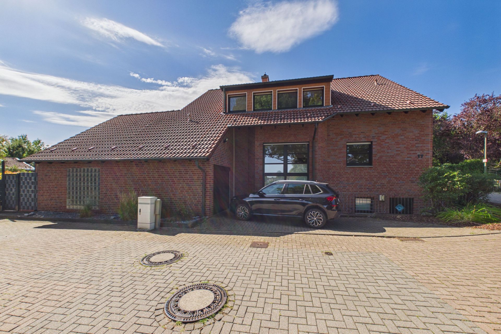 Backsteinwohnhaus mit dunklem Dach und geparktem Auto davor, gepflasterte Straße im Vordergrund.