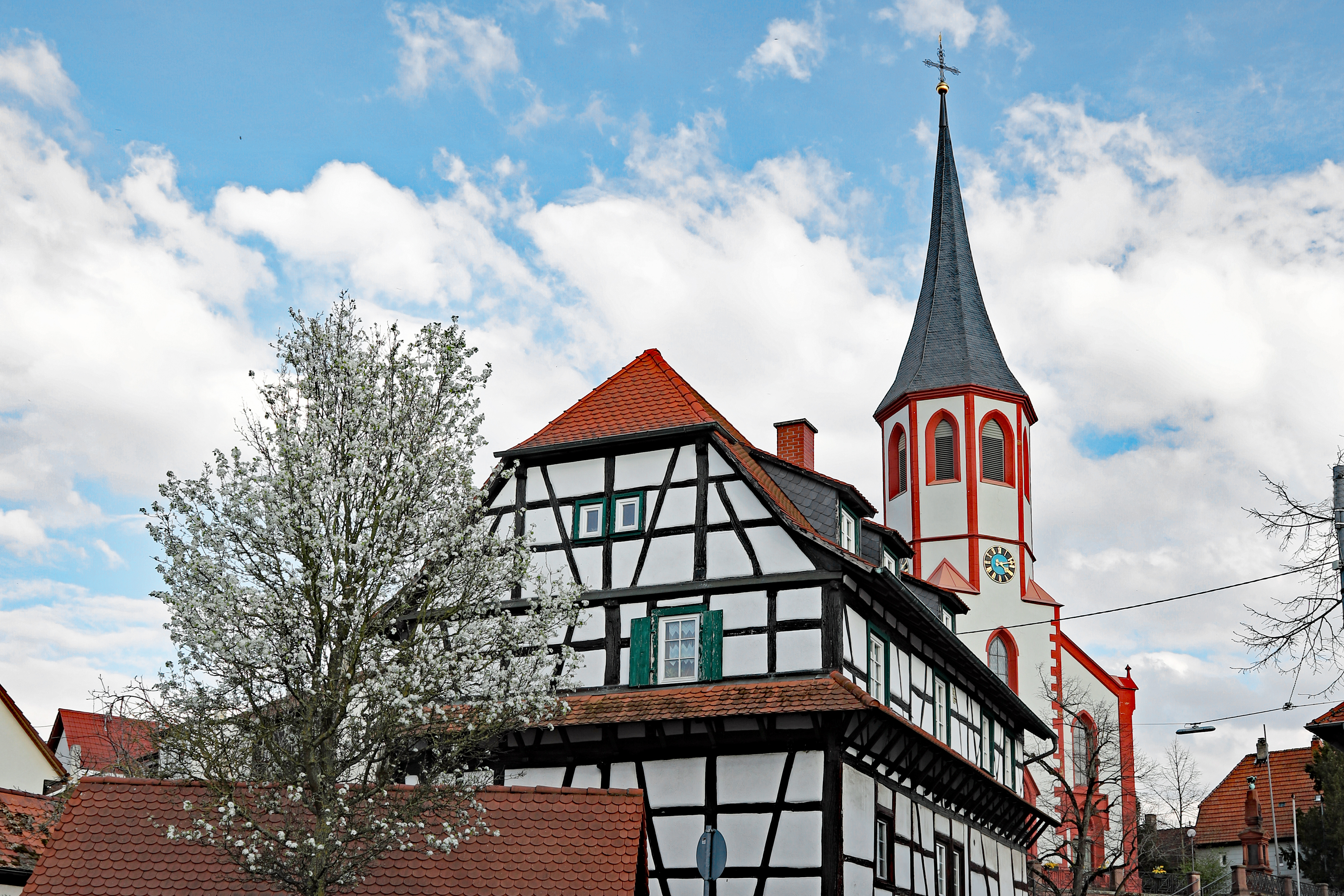 Fachwerkhaus mit rotem Dach, blühender Baum davor, daneben eine Kirche mit hohem Turm.