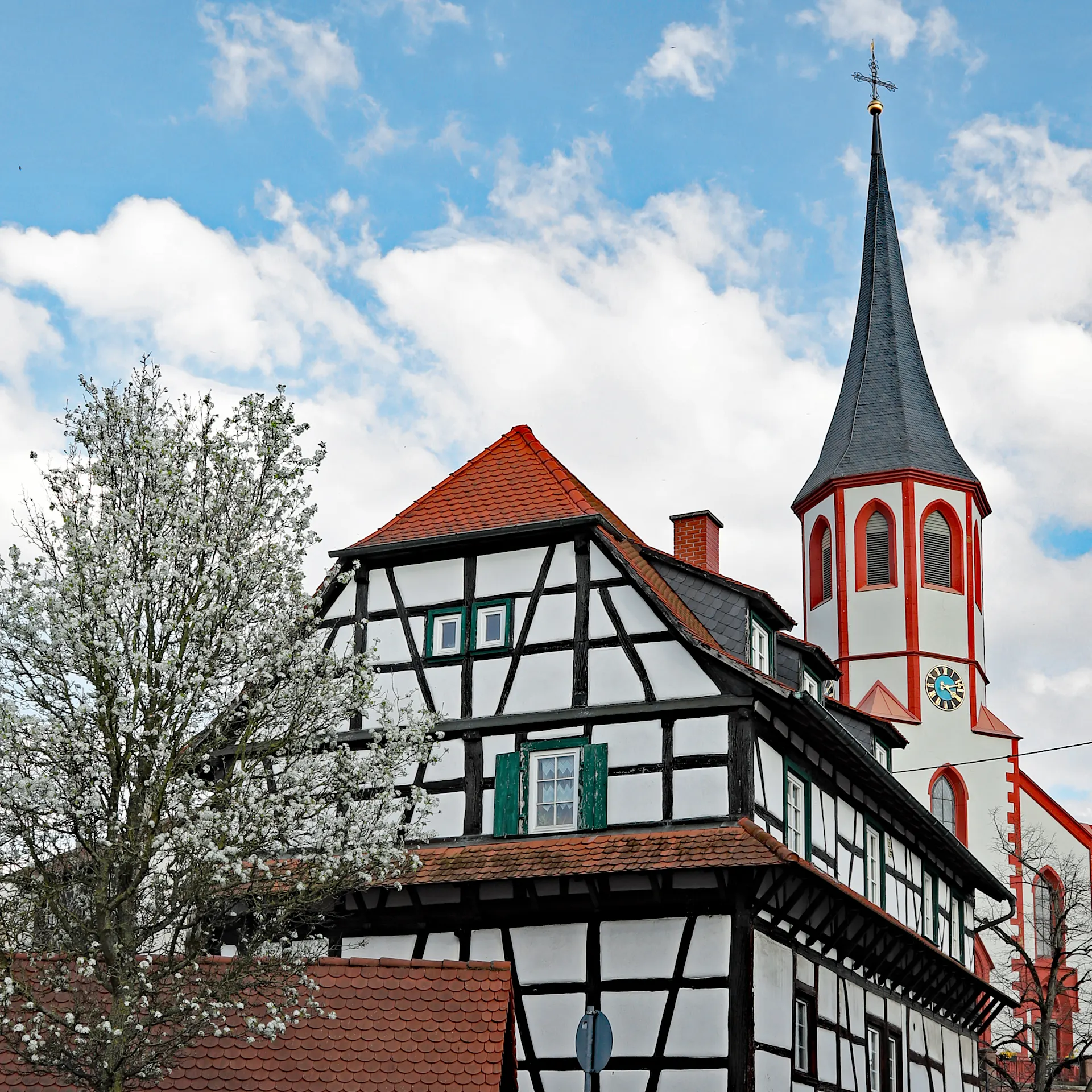 Fachwerkhaus mit rotem Dach, blühender Baum davor, daneben eine Kirche mit hohem Turm.