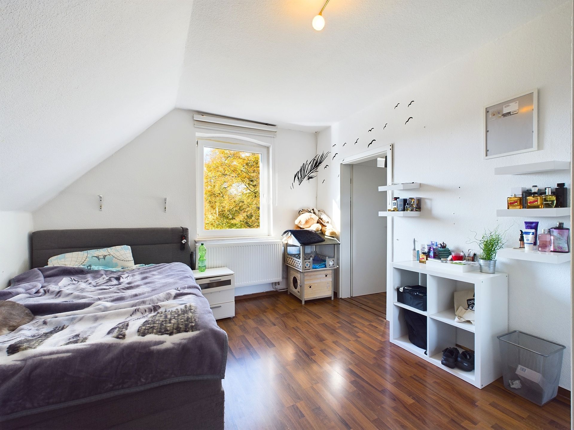 Helles Schlafzimmer mit Bett, Regal, Holzfußboden und Fenster mit Blick ins Grüne.