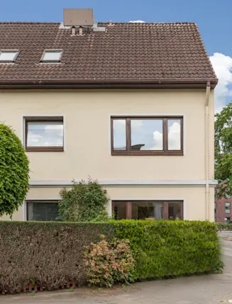 Zweistöckiges Wohnhaus mit braunem Dach, großen Fenstern und gepflegtem Vorgarten an einer Straßenecke.