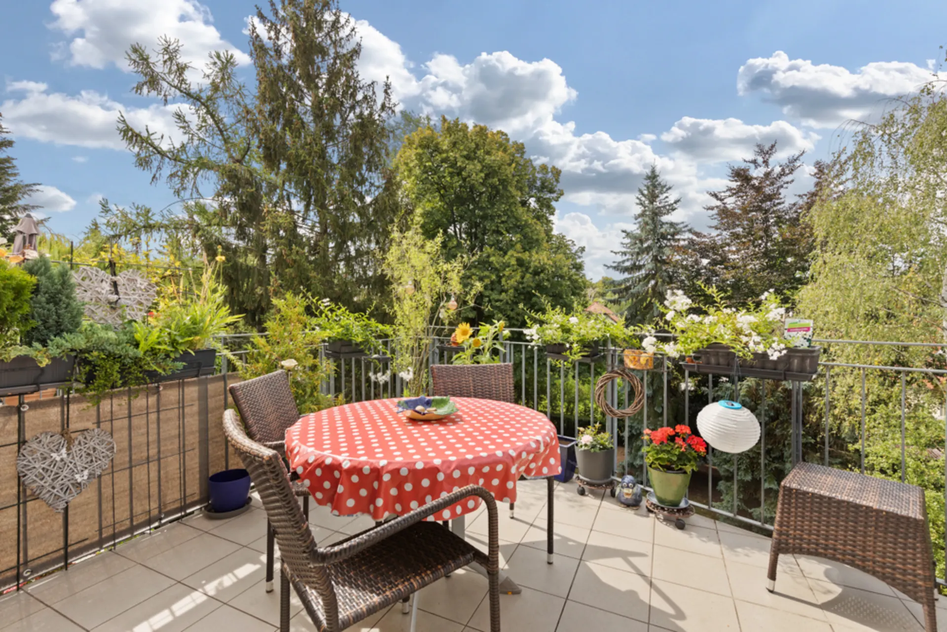Balkon mit Tisch, Stühlen, Pflanzen und Blick auf Bäume bei sonnigem Wetter.