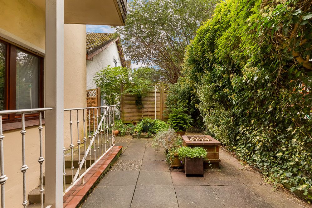 Gepflasterte Terrasse mit Pflanzen, Holztisch und Sichtschutzzaun im Garten eines Wohnhauses.