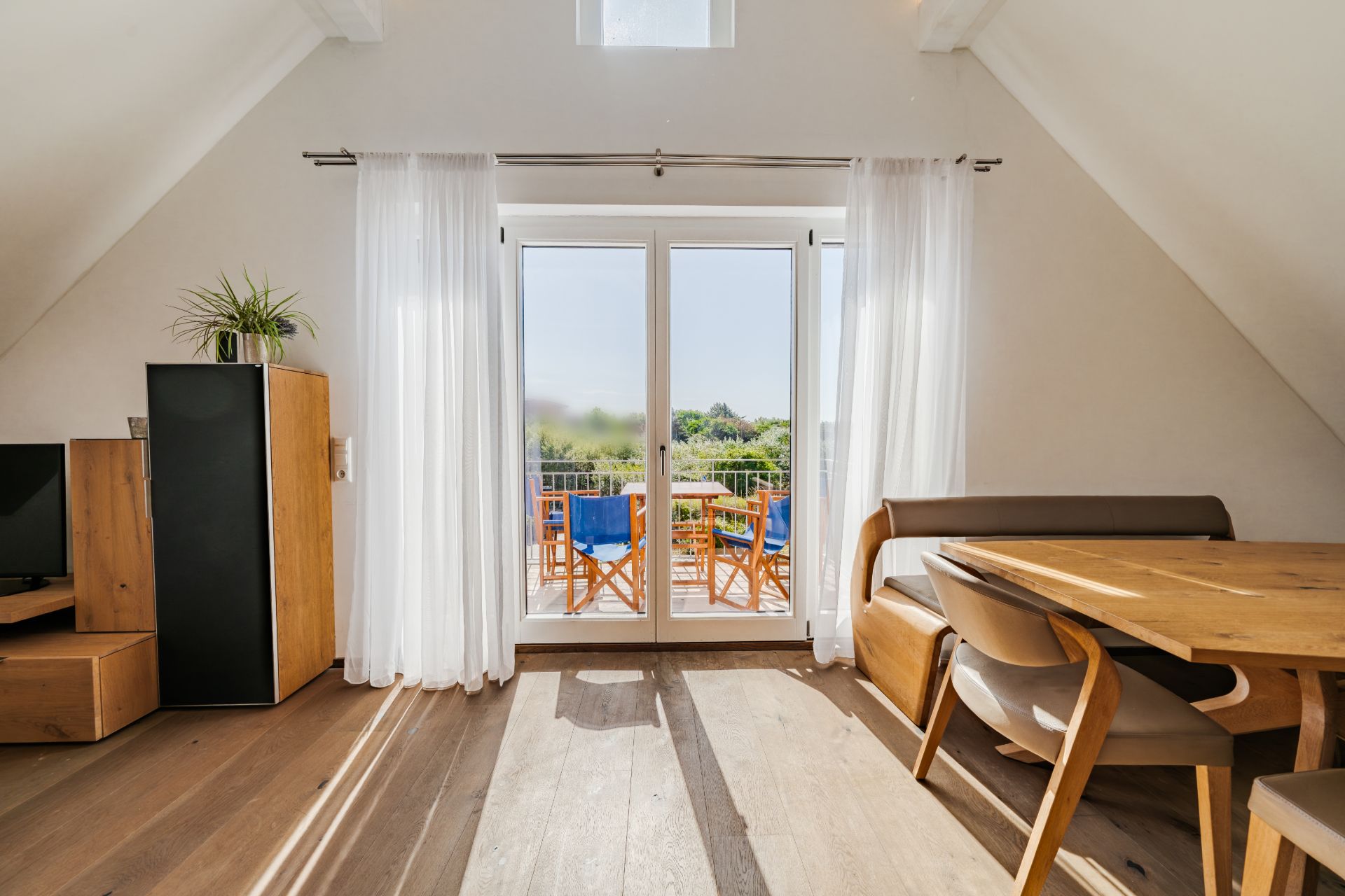 Helles Wohnzimmer mit Essbereich, Holzmöbeln und Zugang zu einem Balkon mit Gartenblick.