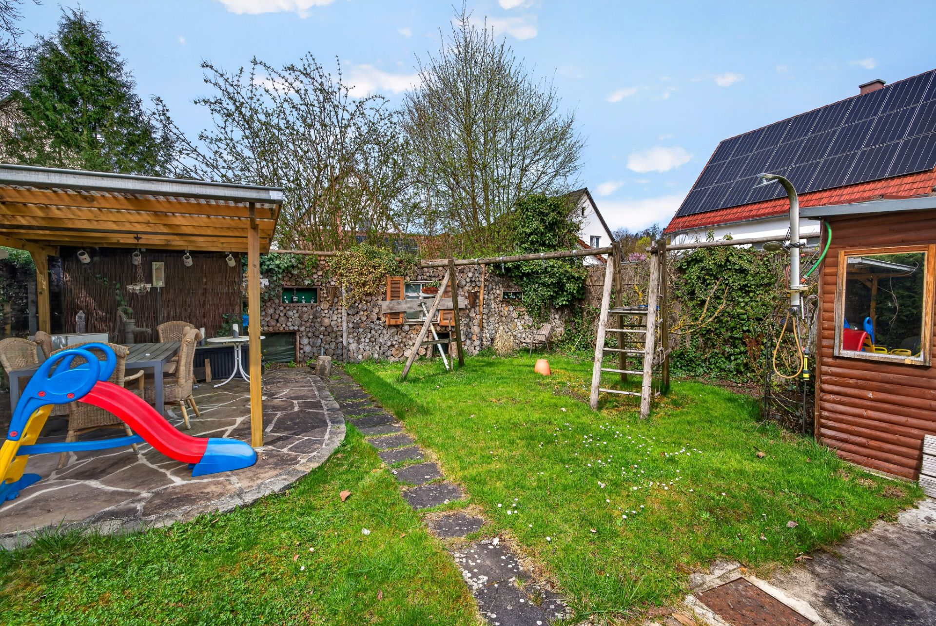 Garten mit Sitzbereich, Rutsche, Schaukel und Haus mit Solarpanels im Hintergrund.