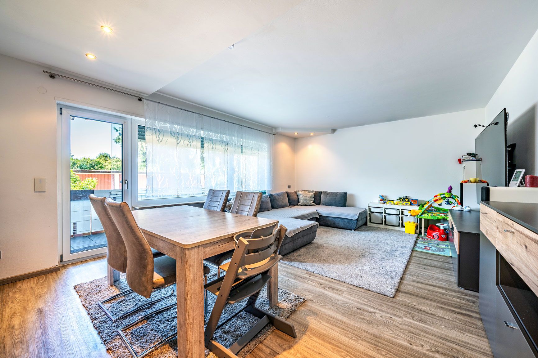 Helles Wohnzimmer mit großer Eckcouch, Esstisch, Kinderstuhl und Spielzeug auf Holzfußboden.