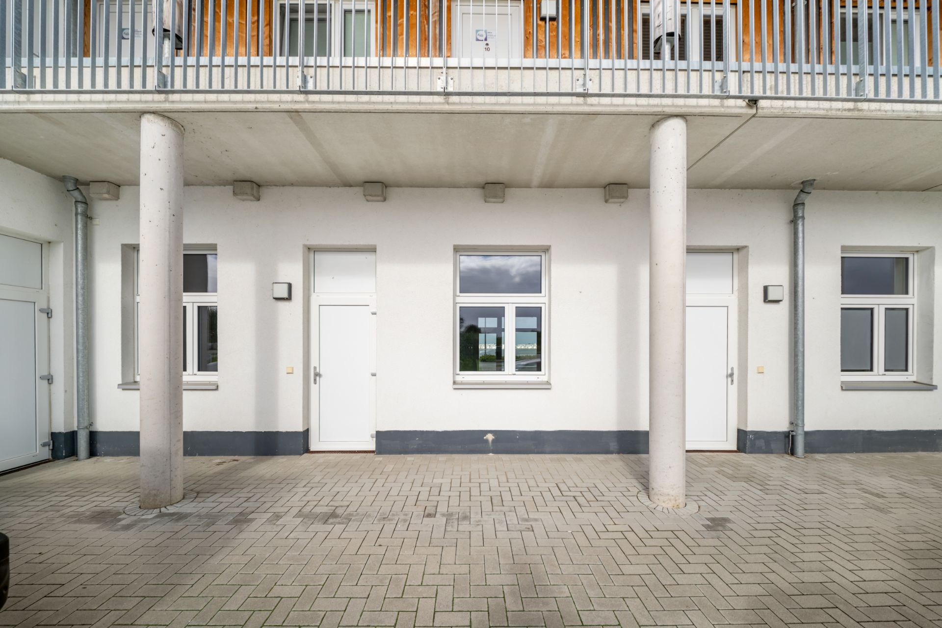 Weiße Hausfassade mit drei Fenstern und zwei Türen, davor Betonpfeiler und gepflasterter Boden.