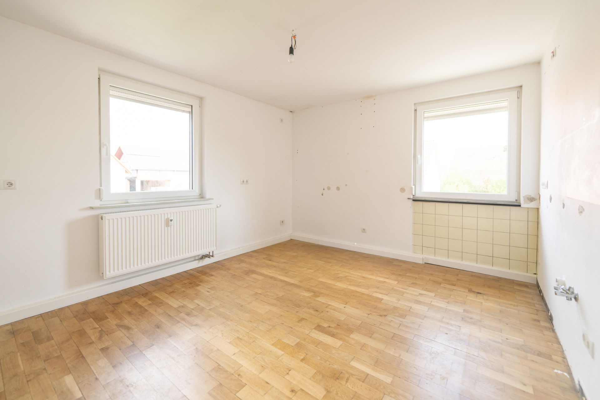 Leerer Raum mit zwei Fenstern, Parkettboden und Heizkörper an der Wand.
