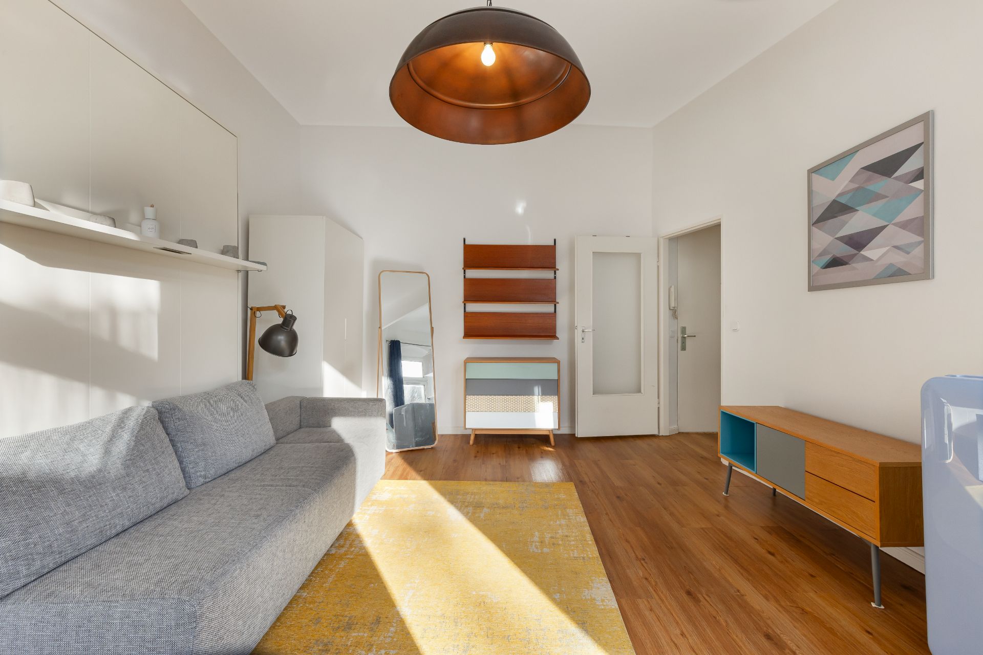 Helles Wohnzimmer mit grauem Sofa, gelbem Teppich, Holzregal und Sideboard.